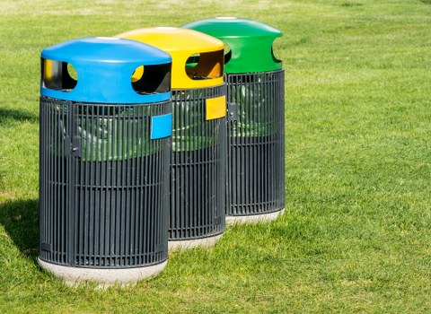 Three cylindrical outdoor rubbish bins with open tops and vertical slats, arranged side by side on a neatly maintained grassy area. The bins are made of black plastic with a textured surface, featuring brightly coloured, rounded domed lids in blue, yellow, and green from left to right. The blue-lidded bin is positioned in the foreground, with the yellow and green-lidded bins slightly behind, creating depth in the image. Each lid has small, semi-circular openings for waste disposal, and the bins are clean and unused, set against a backdrop of vibrant green grass that suggests a residential or commercial outdoor space. The scene appears well-lit, with natural daylight illuminating the objects clearly, emphasizing their weather-resistant finishes. The arrangement and condition of the bins reflect a professional waste management setup in a local Bermondsey environment, aligned with services offered by Business Waste Removal Bermondsey in the London area.