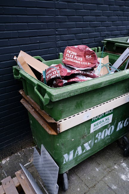 A large, weathered white rubbish bag made of heavy-duty plastic material is placed on a narrow, slightly inclined city street pavement, leaning against a graffitied and stained brick wall with metal s