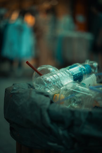 Close-up view of a pile of mixed waste debris behind a black metal fence, consisting mainly of crumpled paper and plastic bags, some with visible printed logos and text. The papers and plastic waste v