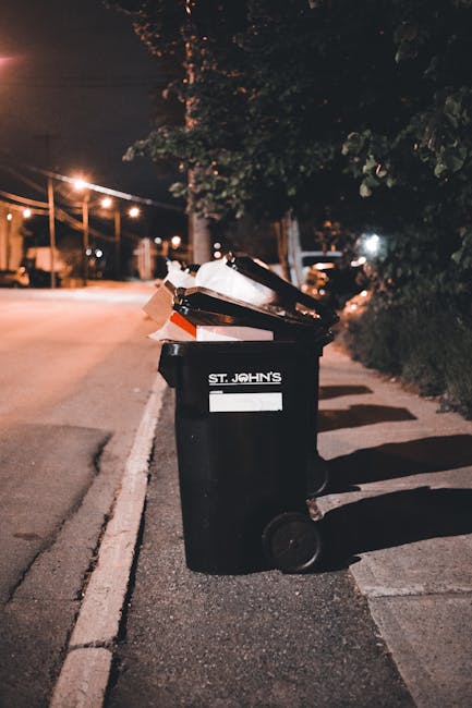 Five large black wheeled rubbish bins with yellow lids are lined up against a plain, light grey wall on a concrete surface. Each bin has a white oval label with black and blue text, positioned central